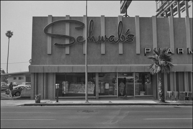 A black-and-white photo features a two-story building with a large façade with no windows on the second story. A large sign on the second story reads “Schwab’s” in cursive.