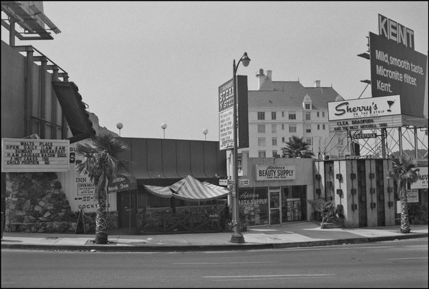 A black-and-white photo features a street scene with several commercial buildings and signage in front of it. In the background is a multi-story building.