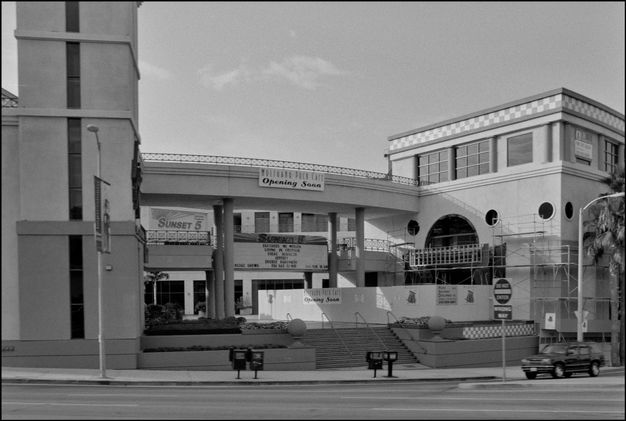 A black-and-white photo features a street scene of a two-story mall complex with a long, uncovered walkway on the second story.