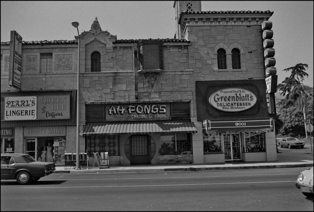 A black-and-white photo features a street scene with a series of two-story commercial buildings that are connected.