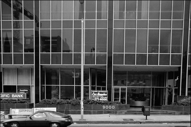 A black-and-white photograph of a building facade with reflective windows. A car is parked out front, and signs for “PACIFIC BANK” and other businesses are visible. The street number “9000” is prominently displayed to the left of the building’s entrance.
