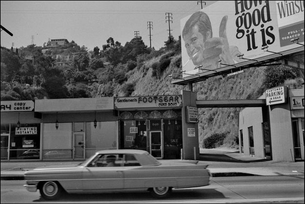 A black-and-white image of a vintage car in motion in front of a strip mall with various stores. A billboard advertising cigarettes with the slogan “How good it is” is at the top right, partially obscured by a tree.