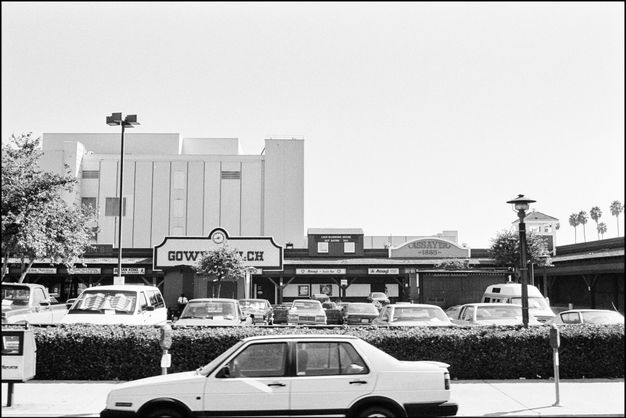 A black-and-white photograph showing a car parked on a street. Behind it are cars parked in a parking lot in front of a single-story strip mall with a sign that reads “Gower Gulch.”