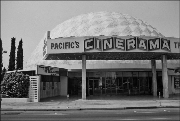 A black and white photo of a movie theater with a white-domed shape roof and large marquee sign that reads “PACIFIC’S CINERAMA TH.” It is flanked by tall narrow trees.