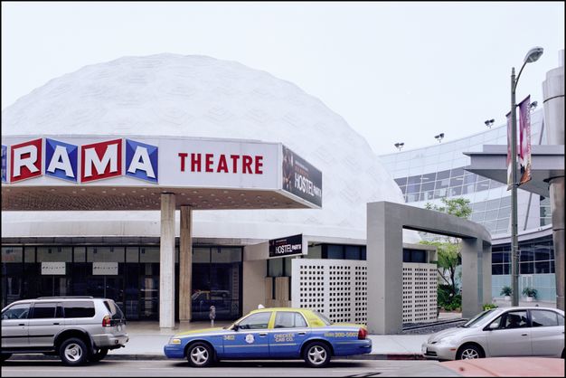 A color photograph of a movie theater with a white dome-shaped roof and large marquee sign that reads “RAMA THEATRE.” A yellow-and-blue taxi is parked in front, and a sign advertising the movie “Hostel Part II” is visible.