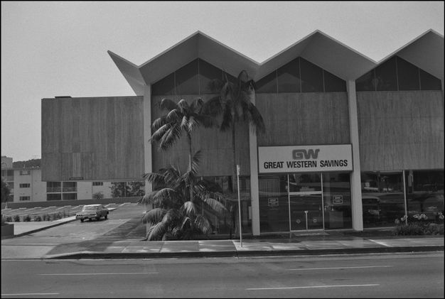 A black-and-white photo of a Great Western Savings bank building with modernist architecture featuring angular rooflines. Palm trees partially obscure the entrance, and a lone car is parked in the adjacent lot.