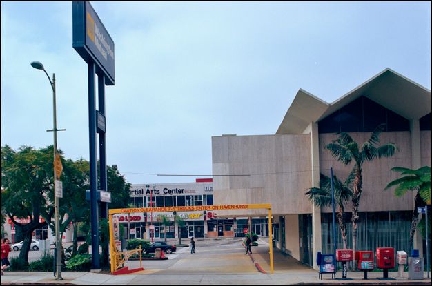 A color photo featuring a mid-century modern building with angular rooflines and large glass windows. A strip mall with various storefronts is visible in the background. The scene includes tall palm trees, colorful newspaper stands, and pedestrians on the sidewalk.