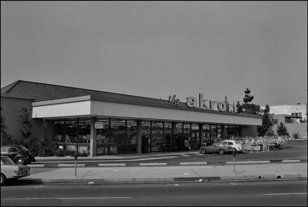 A black-and-white photo features a long, horizontal building with a parking lot in front. A sign on top of the roof overhang reads “the akron.”