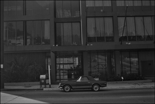 A black-and-white photo features a street scene with a building that is over three stories tall with floor-to-ceiling windows. A single car is parked on the street.