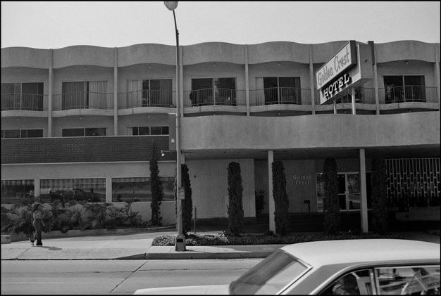A black-and-white photo features a two-story building with a slightly curved repeating roofline and a sign to the right that reads “Golden Crest Hotel.”