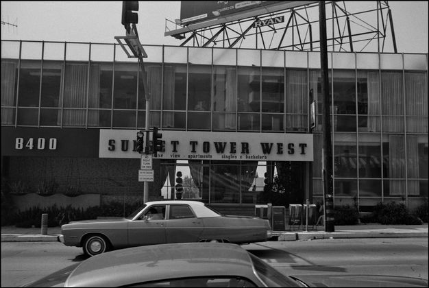 A black-and-white photo features a two-story building with a billboard on top of it. The sign on the front reads “sunset tower west,” and two cars drive by on the street.