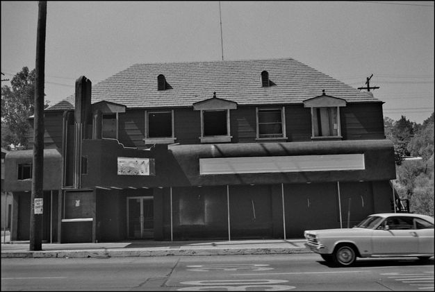 A black-and-white photo features a two-story commercial building at midday, with a sloping, trapezoidal roofline. A car is parked in front of the building at left.