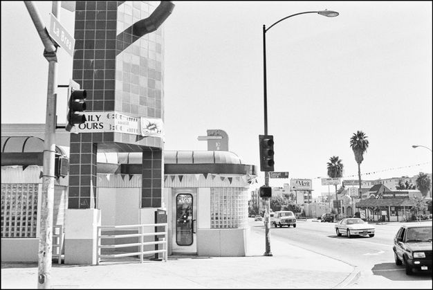 A black-and-white photo features a tiled, two-story building and a tall lamp post. To the right, extending into the background, is a street with multiple cars on it.