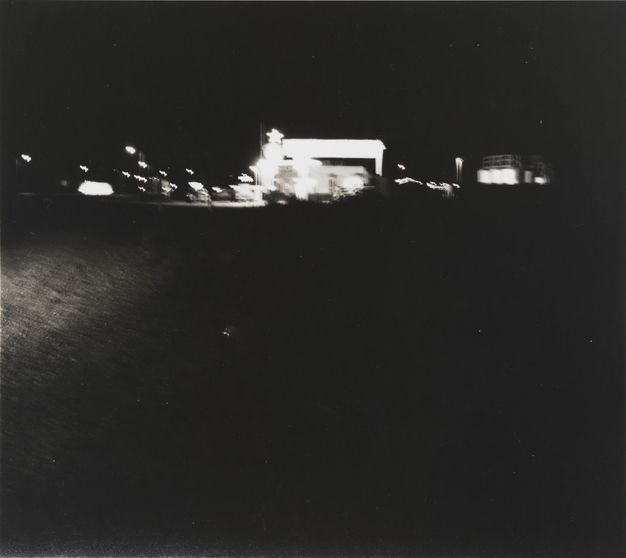 A black-and-white photo of a mostly dark night scene. In the background are blurred lights from what appears to be a town.