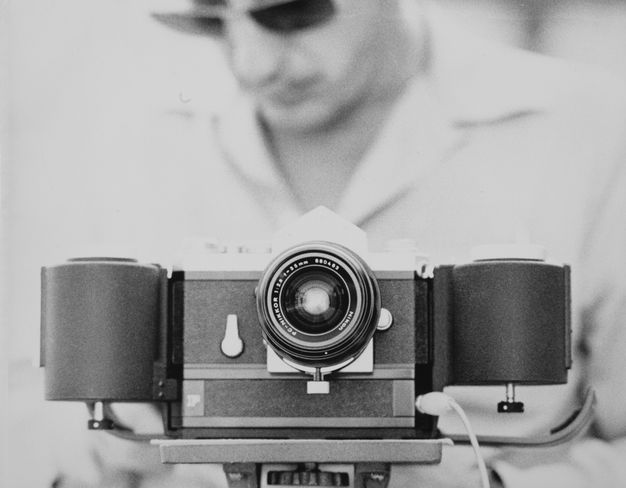 A black-and-white photo features an analog camera in the foreground and, in the background out of focus, a man wearing a white collared shirt and sunglasses looking down.