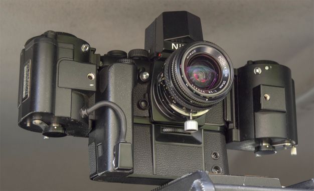 A color photo features a black analog camera with its lens facing forward. Two, white hands hold the bottom and side of the camera.