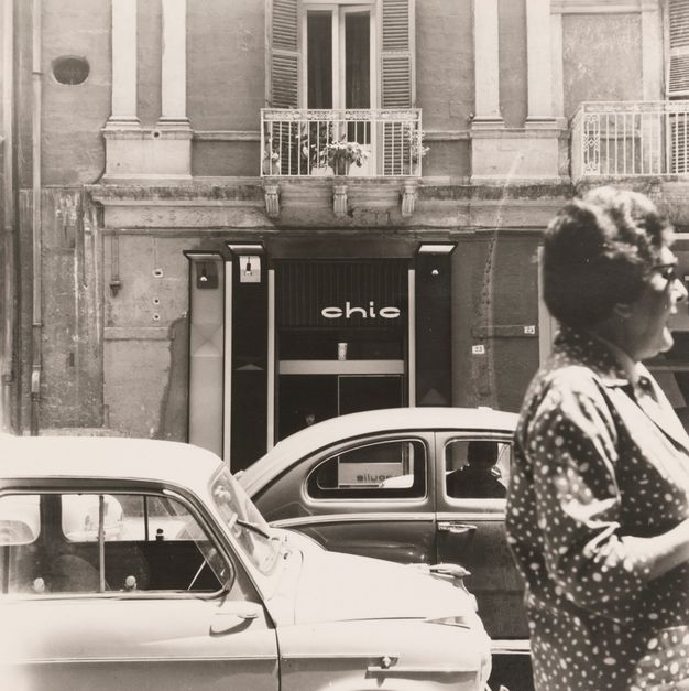 A black-and-white photo showing a bus street scene with two cars and a woman in profile in the foreground. The background is occupied by a two-story apartment building.