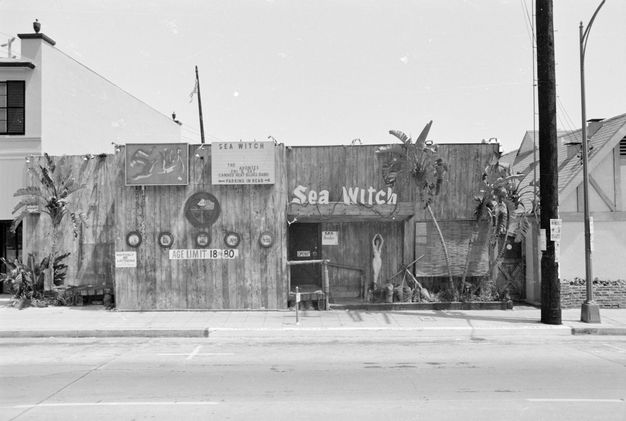 A black-and-white street view showing two one-story buildings flanked by a larger building to the left and a pitched-roofed building to the right. The store front has a sign reading “Sea Witch.”