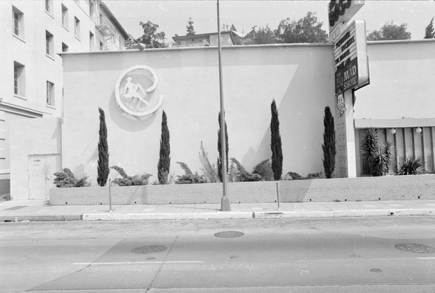 A black-and-white street view of a building with five trees in front, a lamppost bisecting the frame vertically, and large signage attached on tall pole against the building.