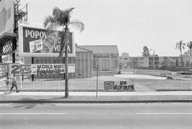 A black-and-white street view showing large billboards and two people walking along the side walk at the left and a building set back from the road with paintage signage reading “Pandora’s Box.”