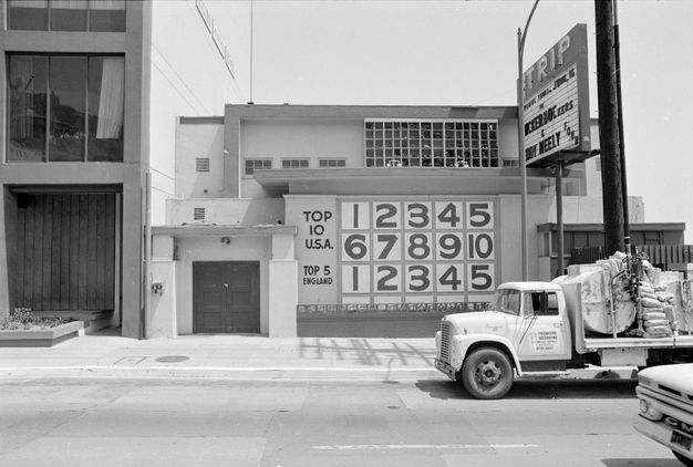 A black-and-white image of a street view with a tall building to the left, a smaller building with numbers painted on the facade, and a small truck bearing a heavy load to the right of the frame.