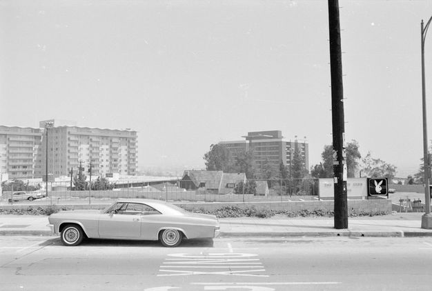 A black-and-white photo of a street view showing a parked car and a wide sidewalk, as well as buildings in the background including high-rise apartment blocks and a pitched-roofed building.