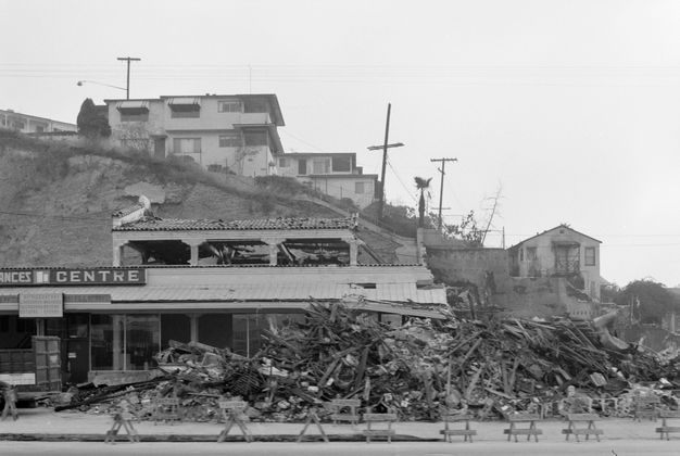 A black-and-white image of a street view showing a damage storefront with collapsed rubble, bricks, and timber, spilling out onto the street. Behind the building a hill extends, with apartment buildings built into it.