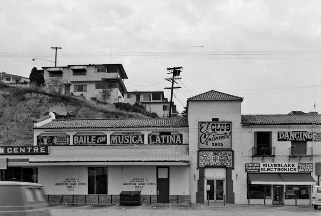 A black-and-white image of a building facade and a hill extended in the background, upon which apartments and telephone wires are placed. The building has a sign reading “El Club Continental.”