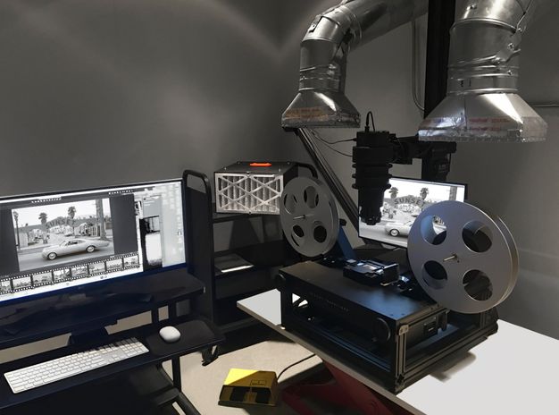 A color photo shows a black machine with a camera mounted in the middle and two film reels on either side. Two silver hoods appear on each side of the machine. Two computer moniters display a Ruscha image of a car.