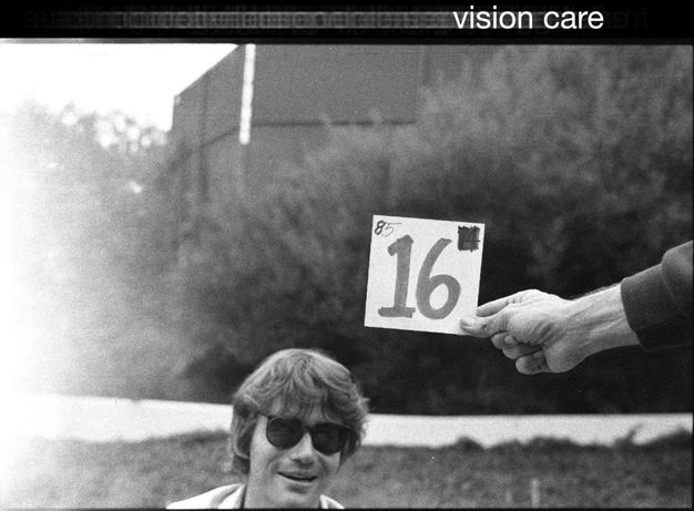 A black-and-white photo shows a man in sunglasses smiling as a hand reaching from out of the frame to the right holds a card that has the number ‘16’ written on it.
