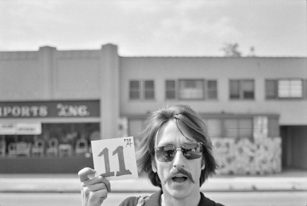A black-and-white photo shows a man with a mustache and sunglasses on a city street holding a square card with the number ‘11’ written on it.