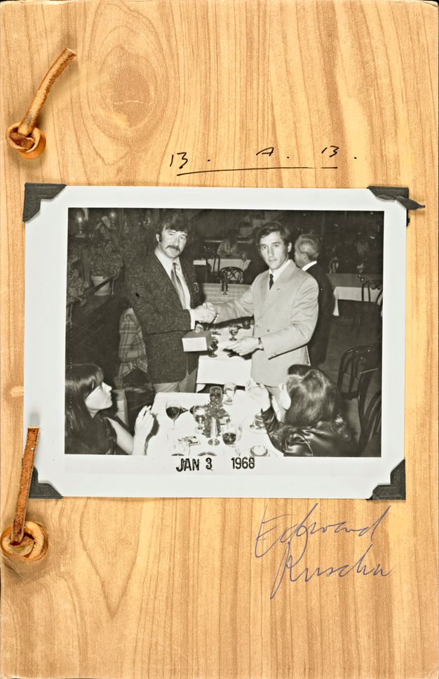 A book cover features a black-and-white photo, set on top of a wood-grained background, of two men in suits shaking hands and two women seated in the foreground looking at them. Cursive on the cover reads “Business Cards” and below the photo “Ed Ruscha.”