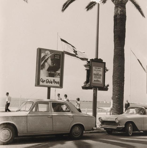 A sepia toned photo shows a street scene, a road with two cars parked, two small billboards, and the ocean in the background. Several people walk beside the ocean.