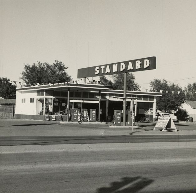 A black-and-white photo features a gas station with the word “Standard” displayed on a sign. There are no cars present and three trees in the background.