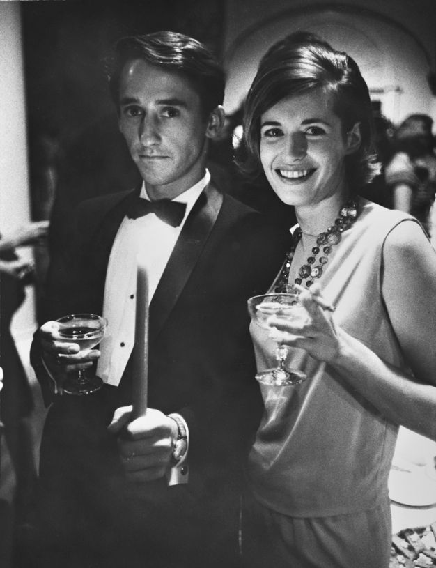 A man in a tuxedo and a woman in evening attire smile and pose together. Each holds a cocktail glass in one hand.