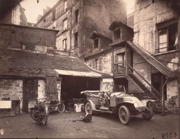 A sepia toned photo features a courtyard between several tall, old buildings. An old car is parked in the foreground.