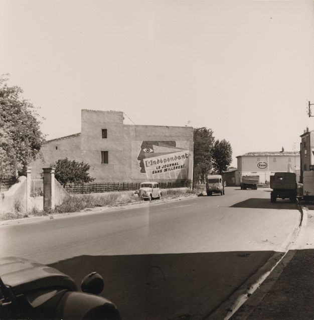 A black-and-white photo features a wide, empty, curved street, shadowed in the foreground, with a two-story building façade in the background.