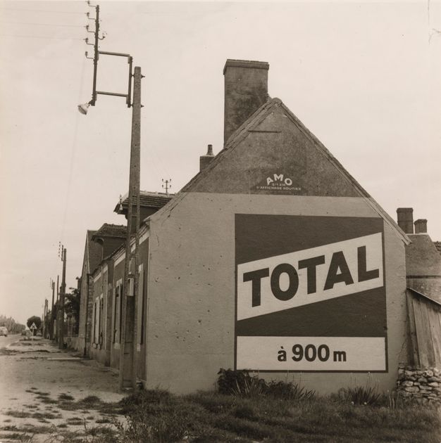 A black-and-white photo shows the side of a building with a peaked roof. On the side is an ad with the word “Total” written in large letters.