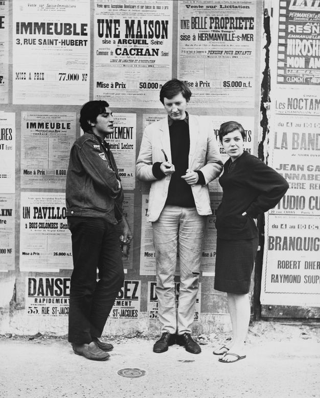 A black-and-white photo features three people standing in front of a wall of posters on a street. The person at right looks towards the camera.