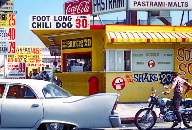 A color photo featuring a bright yellow food stand with many advertisements and signs crowding the foreground and background. A pale gray car and a motorcycle and its rider comprise the foreground.