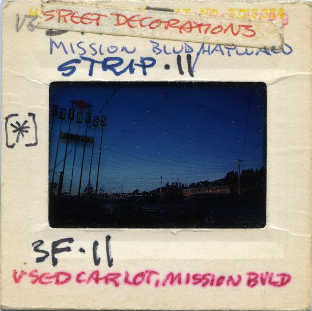 A color photic transparency, in its holder, shows the many strings of illuminated lights over a used car lot with a row of cars for sale. The sign above the lot reads: “Art Bridges South.” The sky is a blue color just before dusk. Handwritten caption by J.B. Jackson reads: “Street decorations/ Mission Boulevard/ Hayward” and “Strip II” and “Used car lot, Mission Boulevard.”