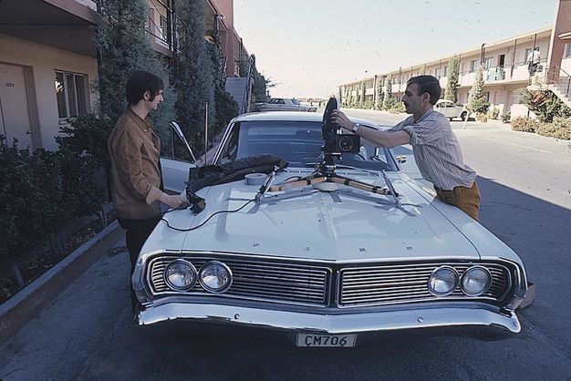 A color photo featuring two men on either side of a white car from the 1960s who are in the parking lot of a motel. The men are tinkering with camera equipment that is placed on the hood of the car.