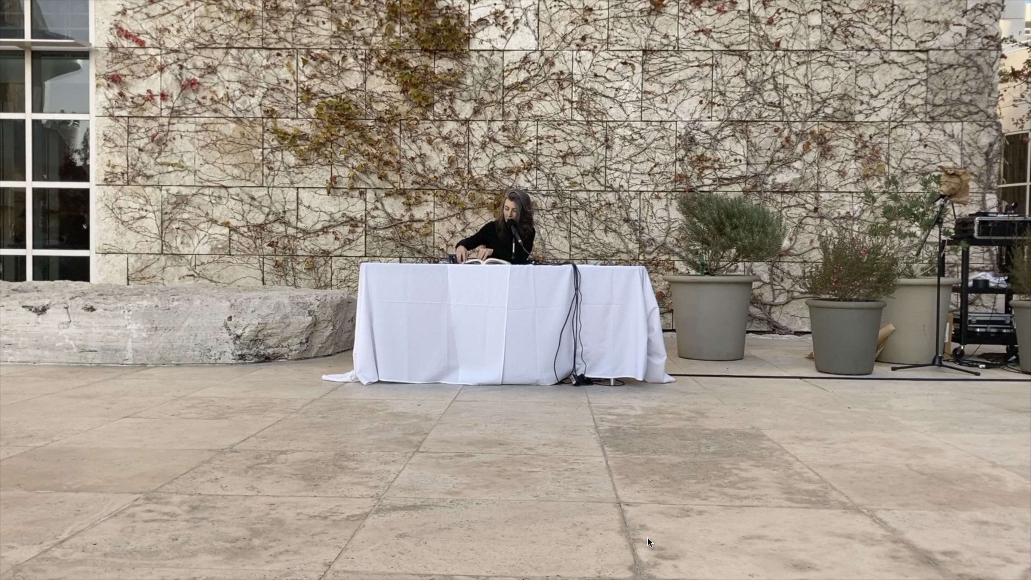 Color video showing middle-aged woman with long, dark hair wearing a black turtleneck sits at a table set up outdoors on museum plaza with a microphone set in front of her on the table. She reads in a brisk, uninflected manner—occasionally whispering—from a book of texts by John Cage.