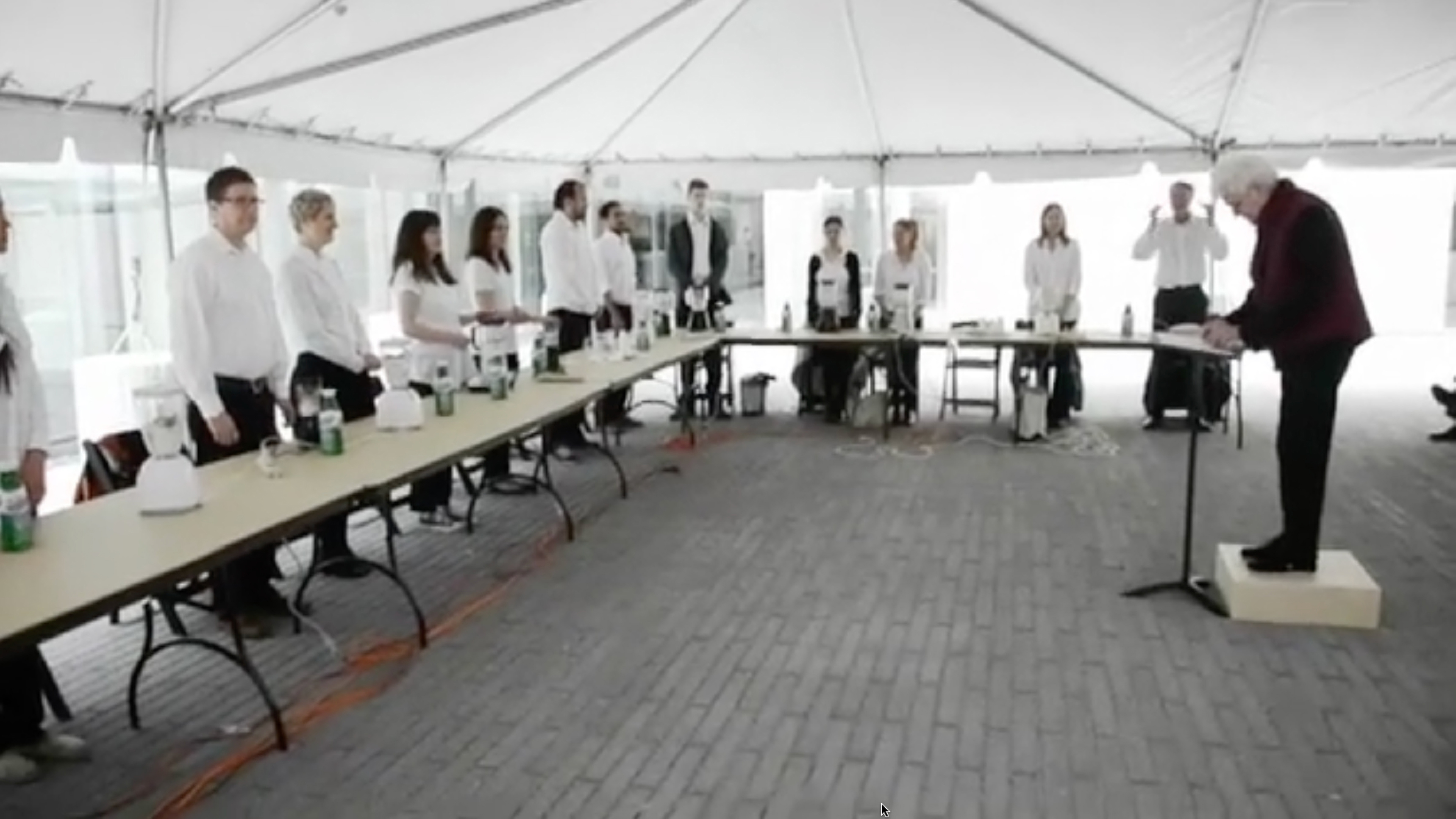 Color video of performance in outdoor tent with performers in white shirts and black pants, each sat at U-shaped table and each with a blender, facing a female conductor (Knowles) at center and an audience behind. Woman first faces away from performers and addresses audience, then conducts performers who turn blenders filled with liquid on or off, pausing halfway through to inspect several blender lids. Knowles returns to her central spot and continues conducting. Finally, Knowles and performers pour the liquid into cups and serve to the audience, some of whom give their opinion on the taste. Video flips between one hand-held camera that follows Knowles and captures close-ups of blenders and hands, and one stable camera behind the audience and facing the table of performers that captures entire scene. 