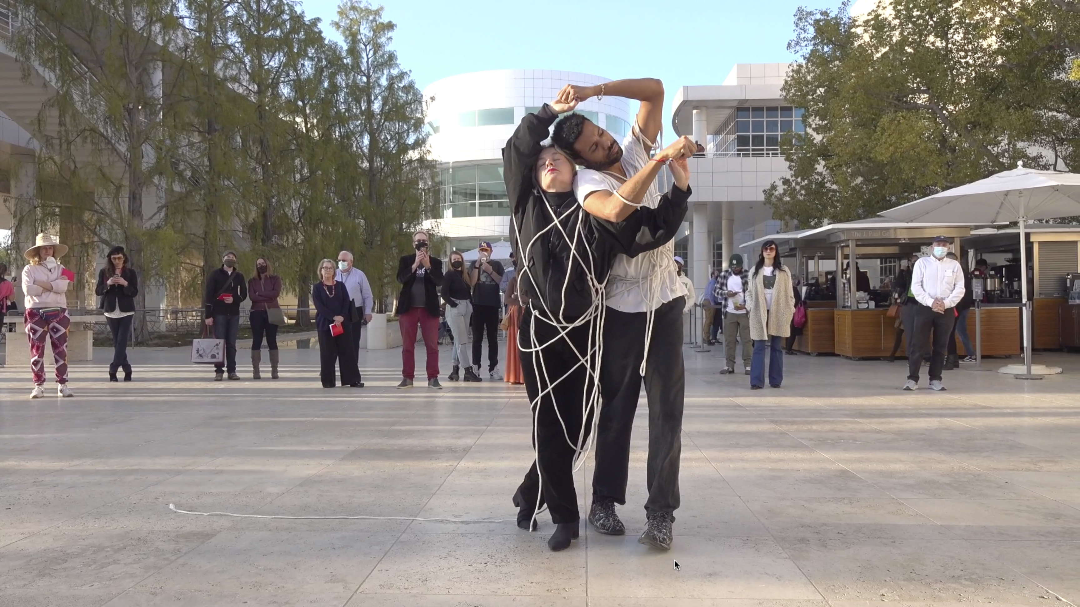 Color video showing woman wearing all black standing outdoors, holding a long white rope that is partially entangled around her, and she uses her arms to entangle her body further. She is approached by a man in white shirt and dark pants who also wears a tangled rope. They touch hands and begin to entwine their bodies while taking various standing and crouching positions while always holding or touching hands. Eventually they spin and then break apart to regard each other from a distance. They crawl on the floor in parallel and then eventually stand up and walk away, holding hands.