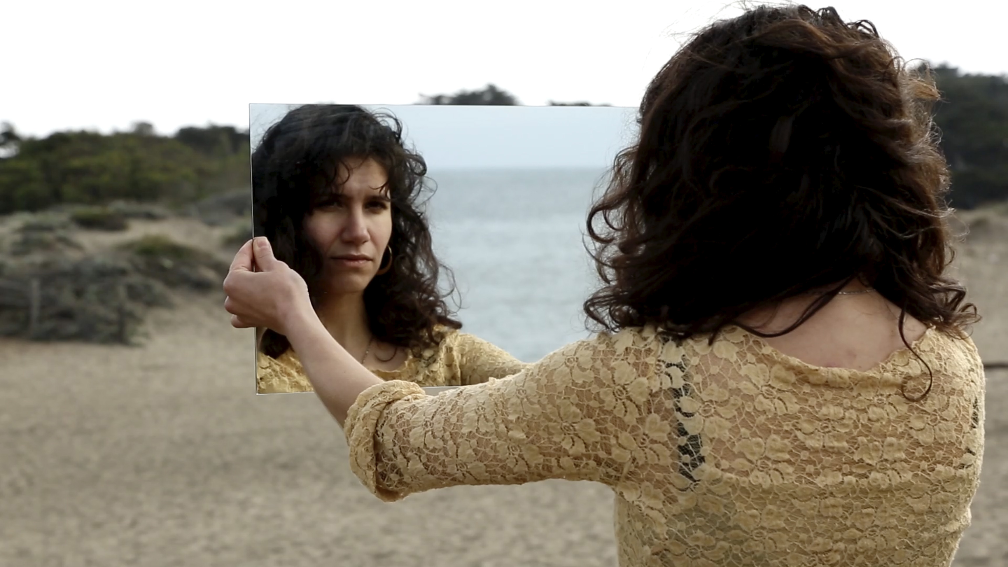 Color video showing sequential, individual episodes of three different women holding and regarding their face in a rectangular piece of mirror as they walk backwards on a beach, into the water. In some moments, the woman’s mirror reveals the cameraman recording her. The women appear to be performing this action on different beaches across the Californian coast. We also see a man, formerly the cameraman recording the women, performing the task while videoing himself with a smartphone enclosed in a plastic bag.