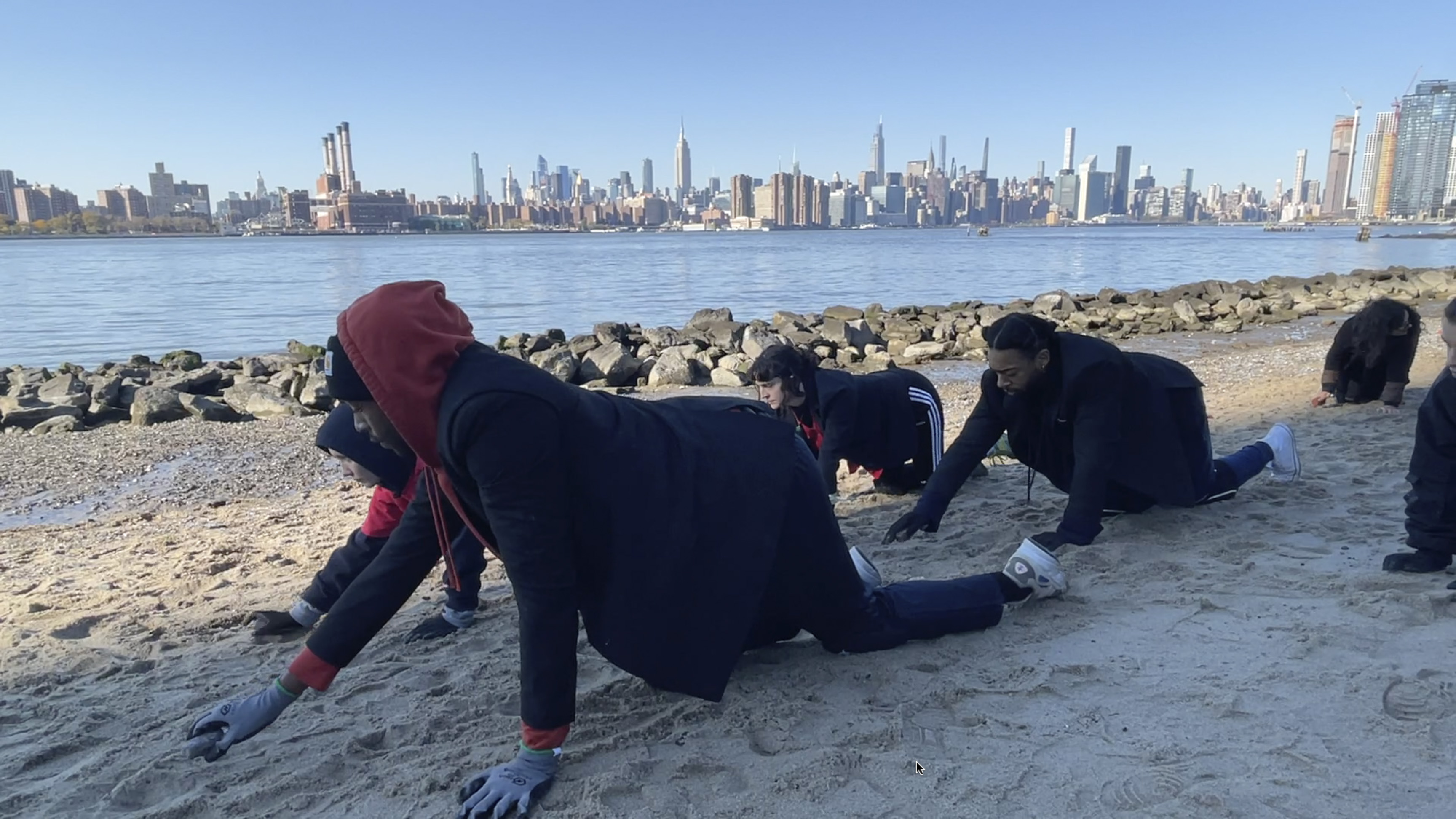Color video showing nine performers wearing warm street clothes and kneepads moving around a rocky beach with the New York City skyline in the background. They collect and compare small rocks, hit rocks together to make sounds, crawl slowly across the sand in unison, throw rocks into the water, rest in small groups, raise their hands toward the sun, and carry one another as they walk away. At one point a woman displays some rocks in her hands as we hear a voice say, “Well that’s new isn’t it?” The soundtrack is a wandering landscape of synthesizer tones that are mostly harmonious but with some moments of dissonance.