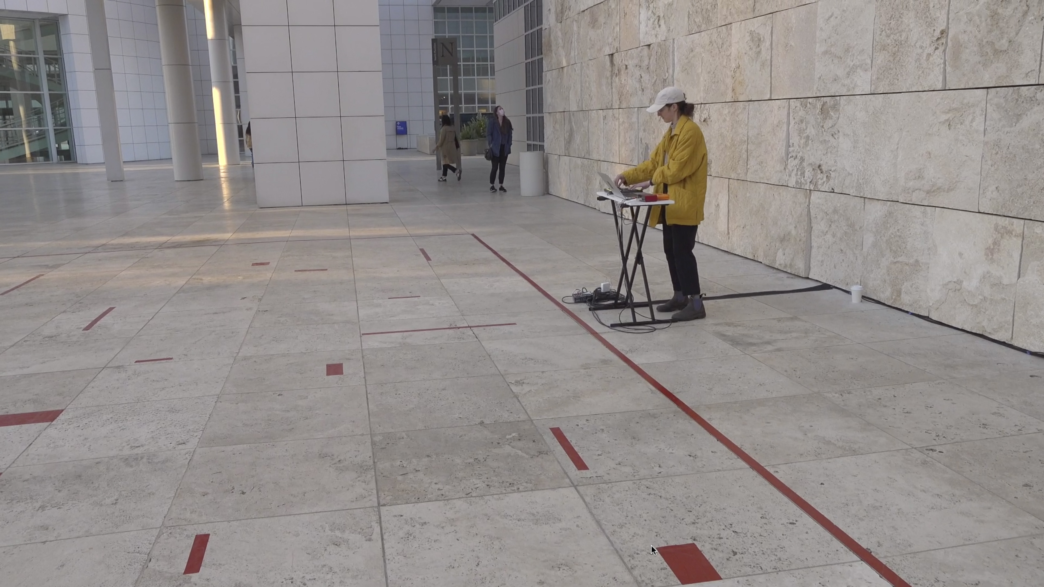 Color video showing a woman wearing a yellow jacket and white baseball cap standing outdoors on a museum plaza, and positioned at the back of a square performance area demarcated by lines of red tape on the ground. Inside the square area appear randomly placed shorter lengths of red tape positioned perpendicularly to one another. In front of the woman is a small table with a laptop and mixing board on it. She uses her hands to manipulate the electronics, which make an abstract soundscape of clicks, beeps, and brief drones that sound like aircraft passing by. At one point, three children run into the performance area to get a closer look.
