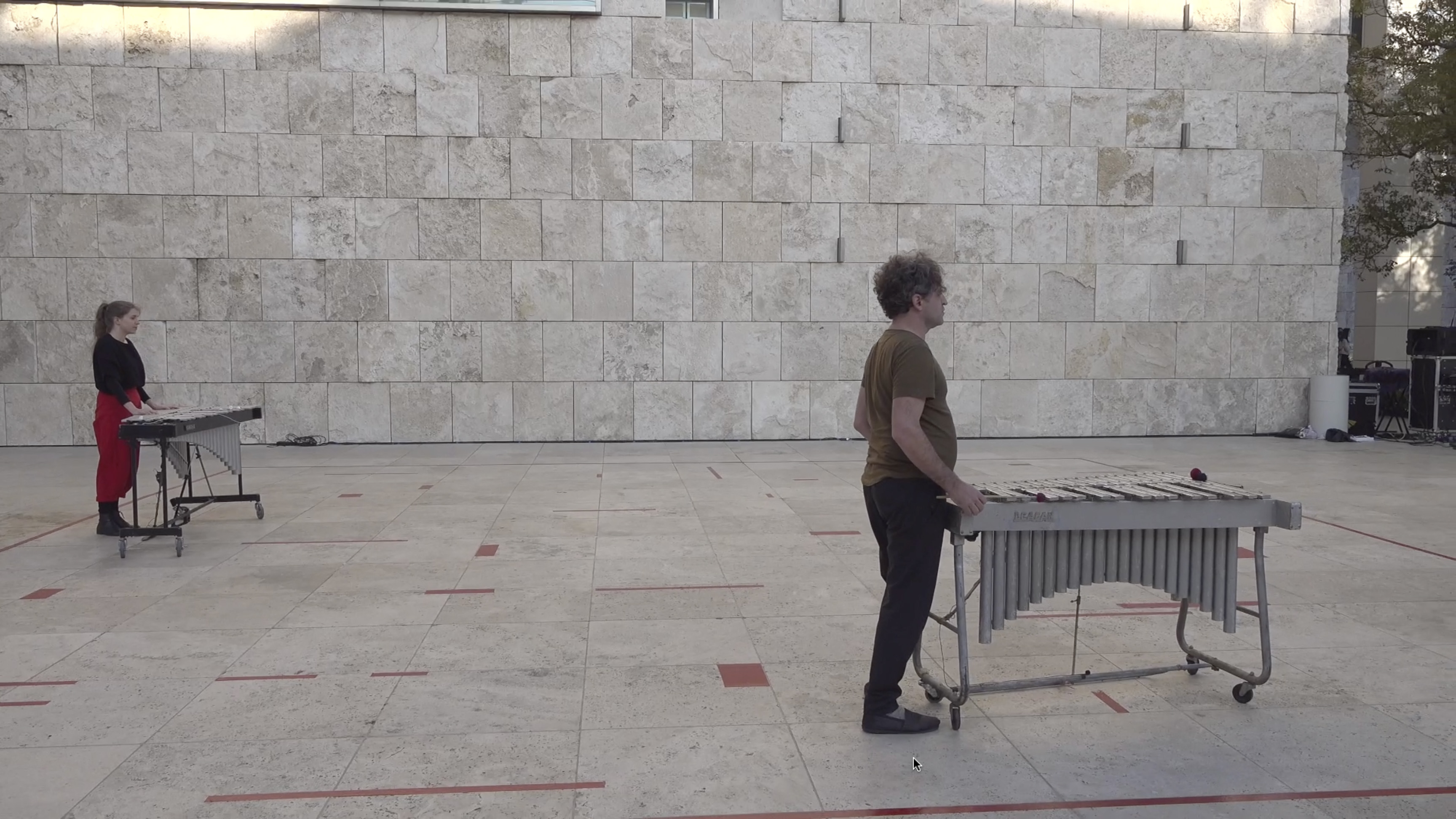 Color video showing a man and a woman wearing street clothes positioned within a square performance area demarcated by lines of red tape on the ground. Inside the square area appear randomly placed shorter lengths of red tape positioned perpendicularly to one another. The performers each have a vibraphone on wheels that they push around the performance area and with which they make spare, occasional tones. The woman sometimes makes abstract sustained tones with her voice, walks around the performance area, and lies down on the ground and on top of the xylophone. The xylophones’ wheels make scraping sounds against the ground as they are being moved.