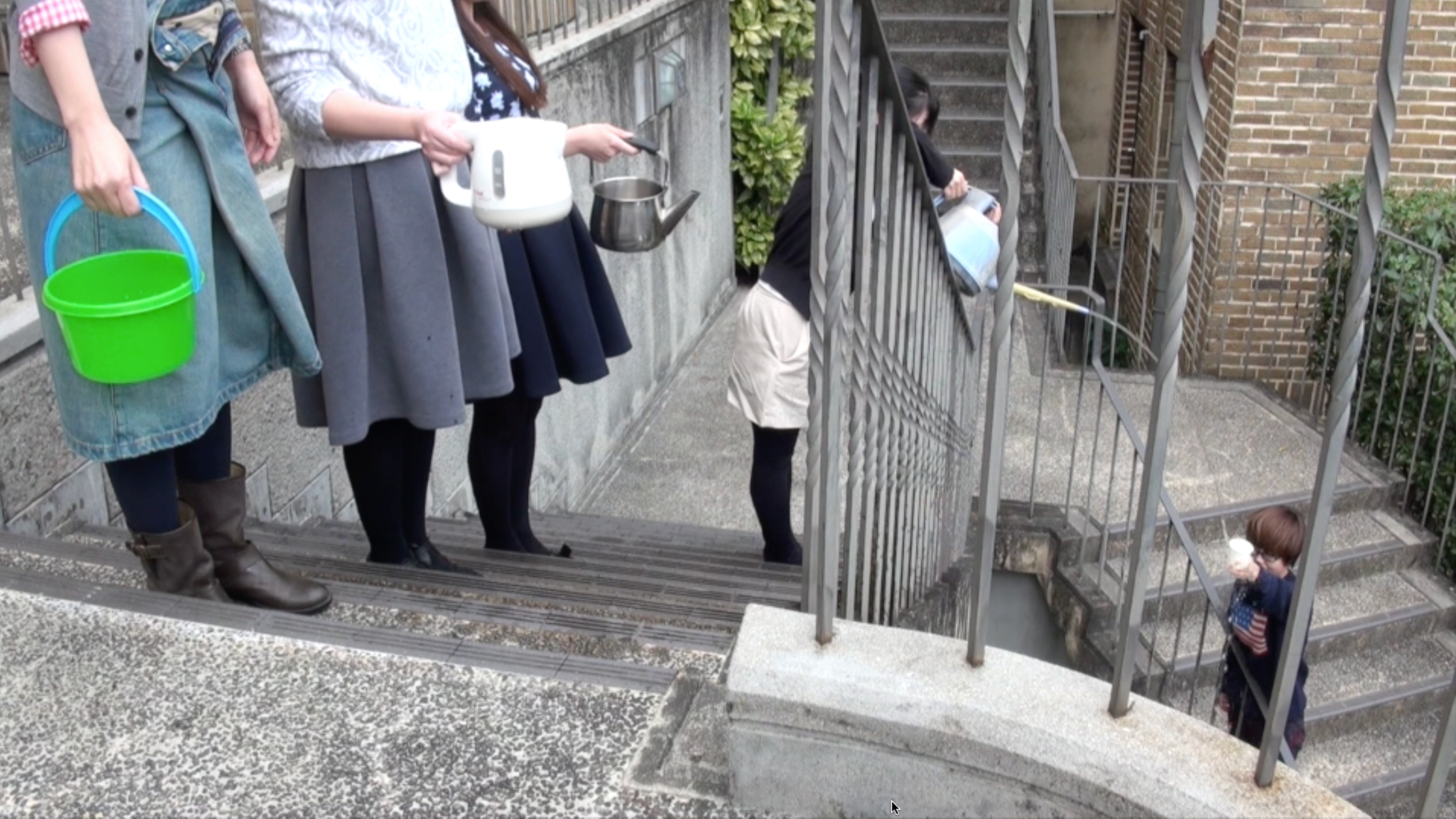 Fixed frame color video of five people standing outside on a concrete staircase, each holding a different vessel filled with water. Each person in succession pours the contents of their vessel into the vessel of the person on their left, from bucket to plastic kettle to metal kettle to watering can to paper cup, during which, water spills onto the staircase. Once the watering can has poured its entire contents into the cup, the video ends.